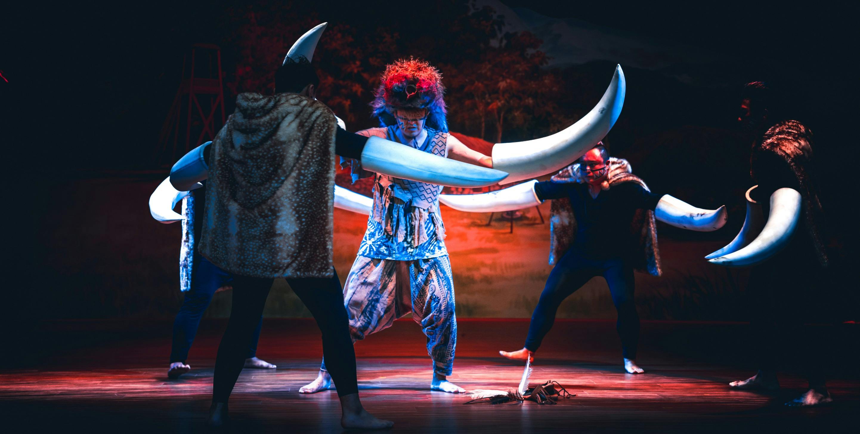Performers in a theatrical scene, one wearing a costume with large tusks and others in animal-themed attire on stage.