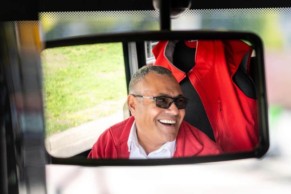 A man with short hair, wearing glasses and a red jacket, is smiling, seen through a vehicle's rearview mirror.