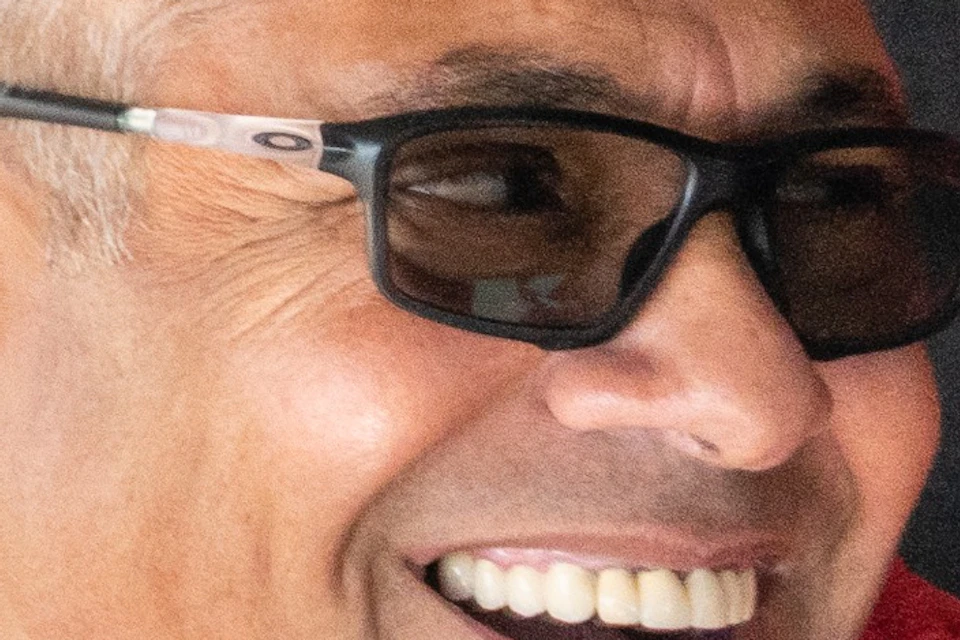 A man with short hair, wearing glasses and a red jacket, is smiling, seen through a vehicle's rearview mirror.