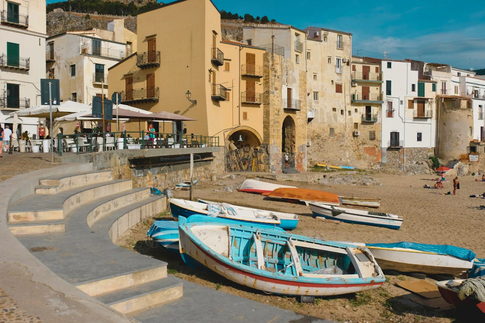 Beached fishing boats on a sandy shore with old buildings and an outdoor café in the background under a clear blue sky.