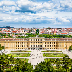 Ett stort gult palats med tre våningar, formella trädgårdar och en stadsbild i bakgrunden under en molnig himmel.