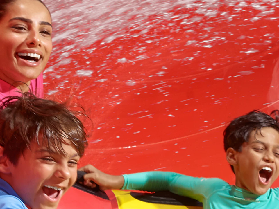 Four people in a yellow and red raft slide down a winding red water slide, joyfully screaming and holding on tightly.