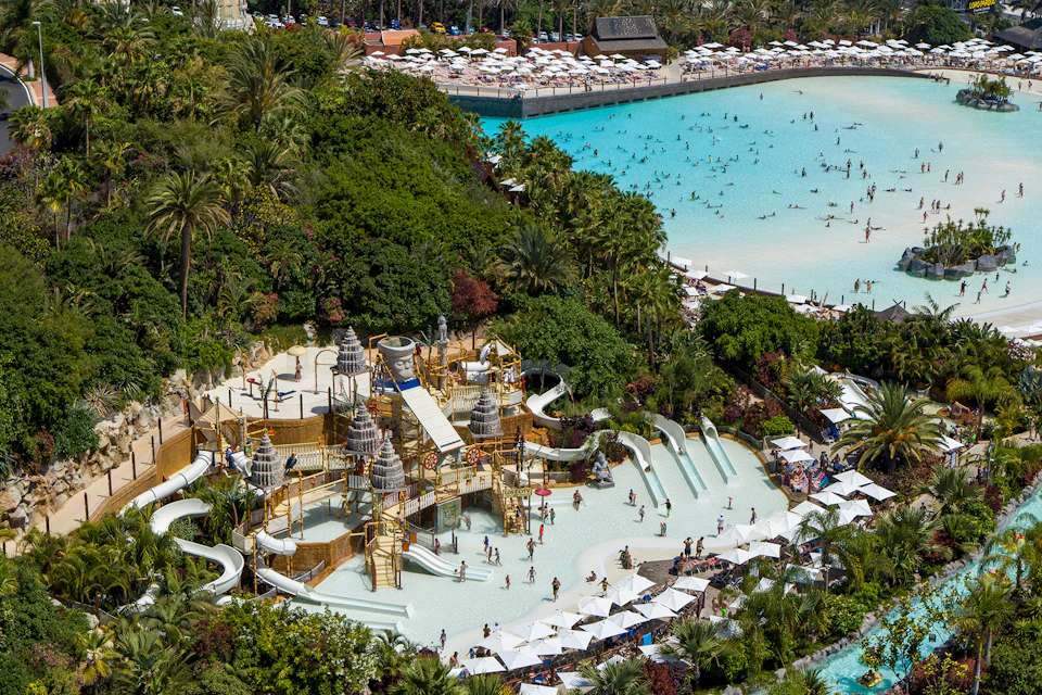 Aerial view of a water park with elaborate slides, pools, and lounging areas; a large pool with numerous swimmers in the background.