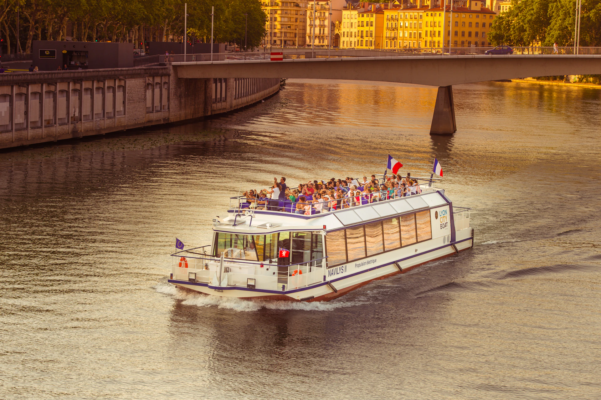 A white river cruise boat with passengers, flags, and a bridge in the background on a calm waterway at sunset.
