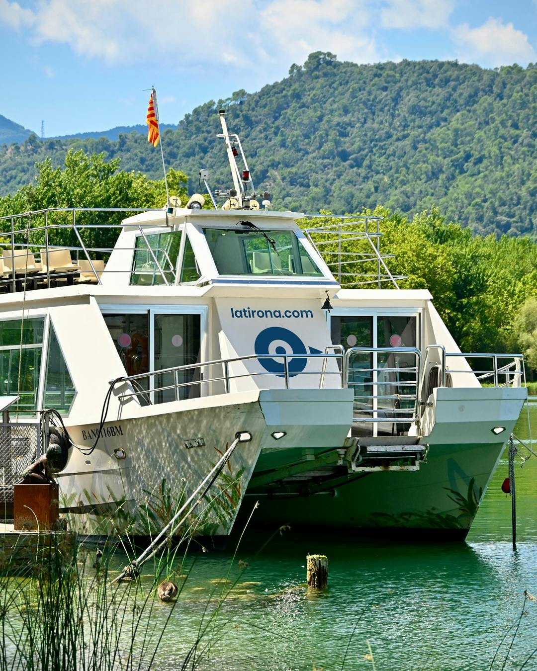 Un catamarà blanc amb el text "latirona.com" va atracar a prop d'un folre exuberant i verd contra un teló de fons de turons boscosos.