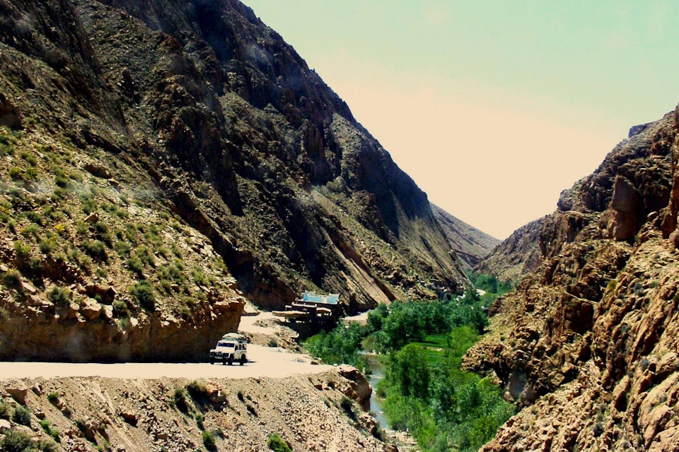 A narrow road winds through arid mountains, a white vehicle parked roadside, with green foliage and a small creek below.