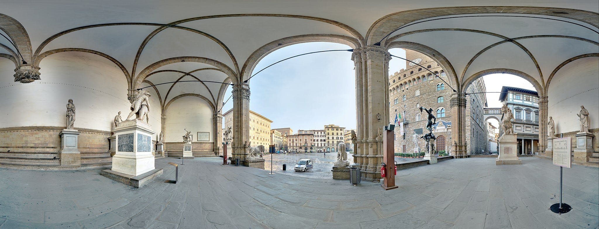 Covered outdoor gallery with stone archways, several classical statues, and a view of surrounding historic buildings.