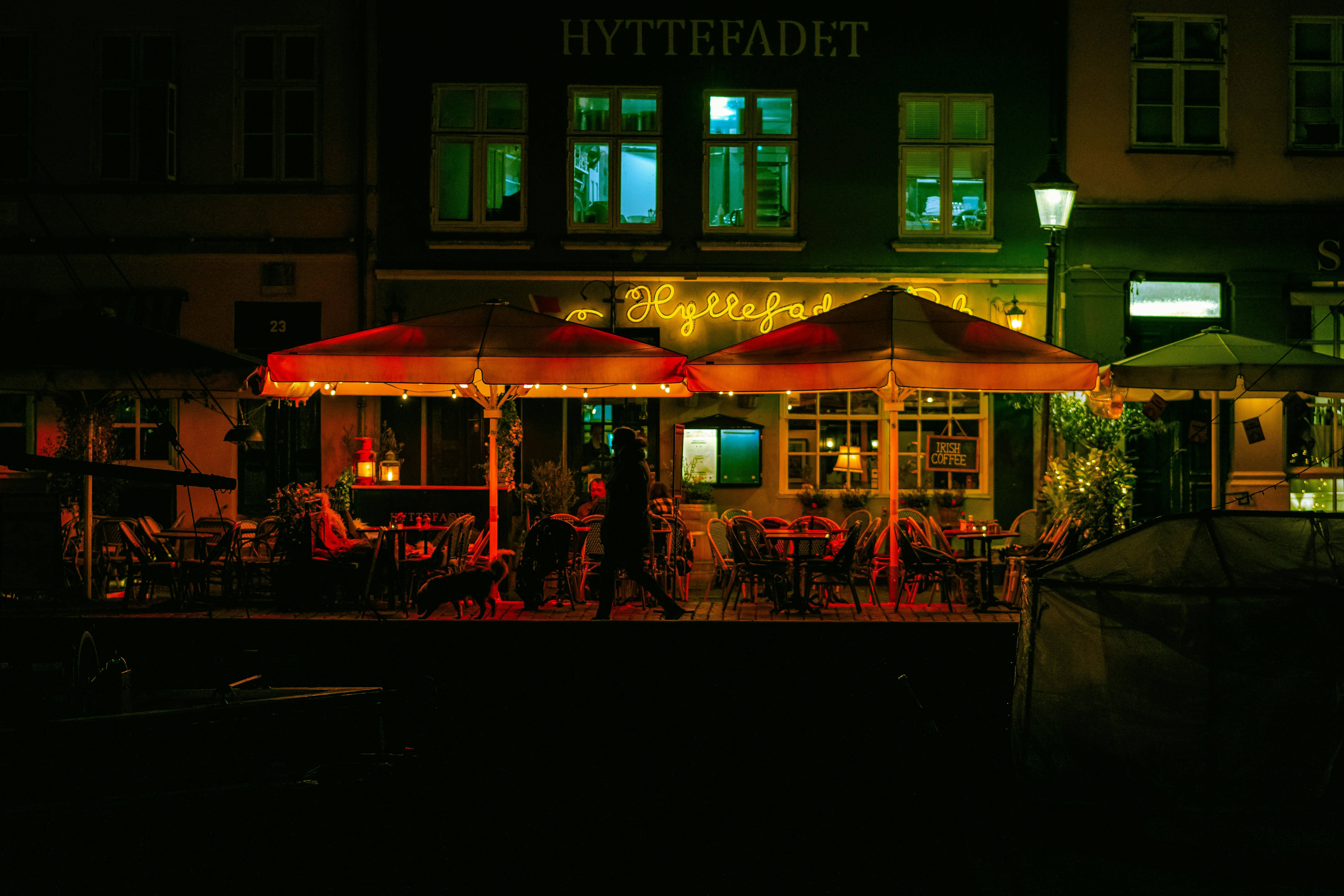 Venkovní kavárna v noci s červenými deštníky a neonovými nápisy, siluetami lidí a psů, slabě osvětlenými stoly a židlemi.