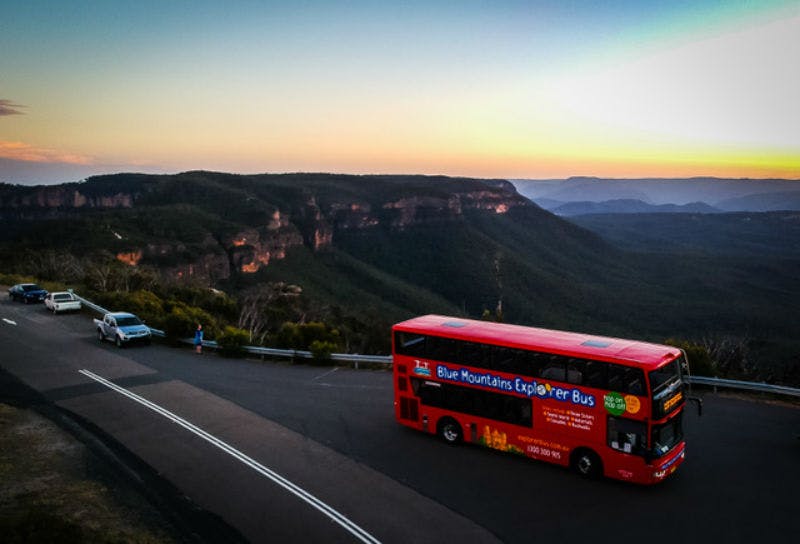 A red double-decker bus labeled "Blue Mountains Explorer Bus" on a mountain road overlooks a vast forested valley at sunset.