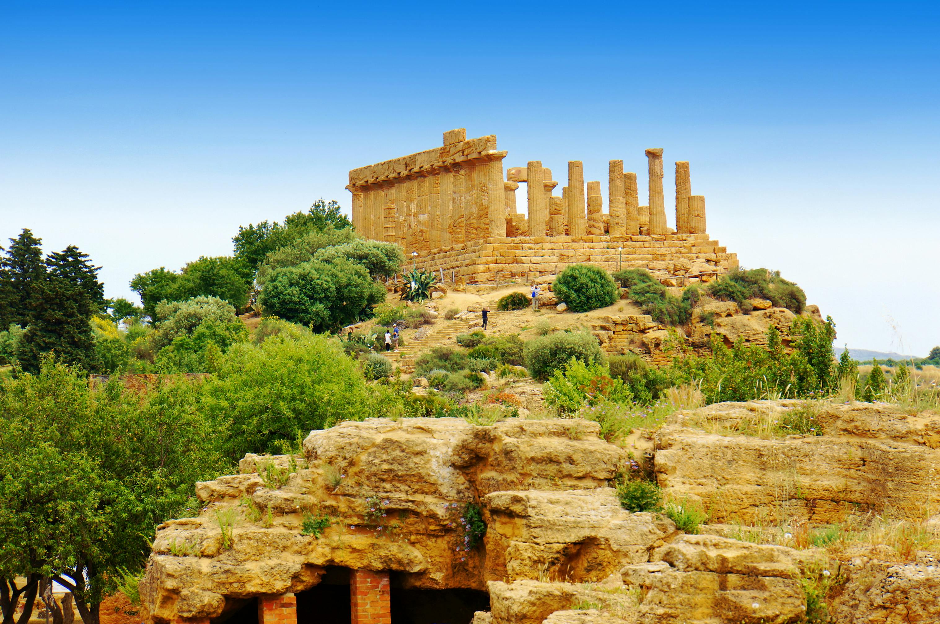 Ancient temple ruins with intact columns and a partially standing structure, surrounded by greenery under a clear blue sky.