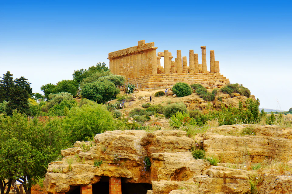 Ancient temple ruins with intact columns and a partially standing structure, surrounded by greenery under a clear blue sky.