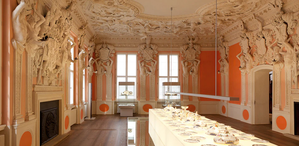 A grand room with ornate, sculpted walls and ceiling, large windows, and a well-lit display of fine porcelain on a central table.