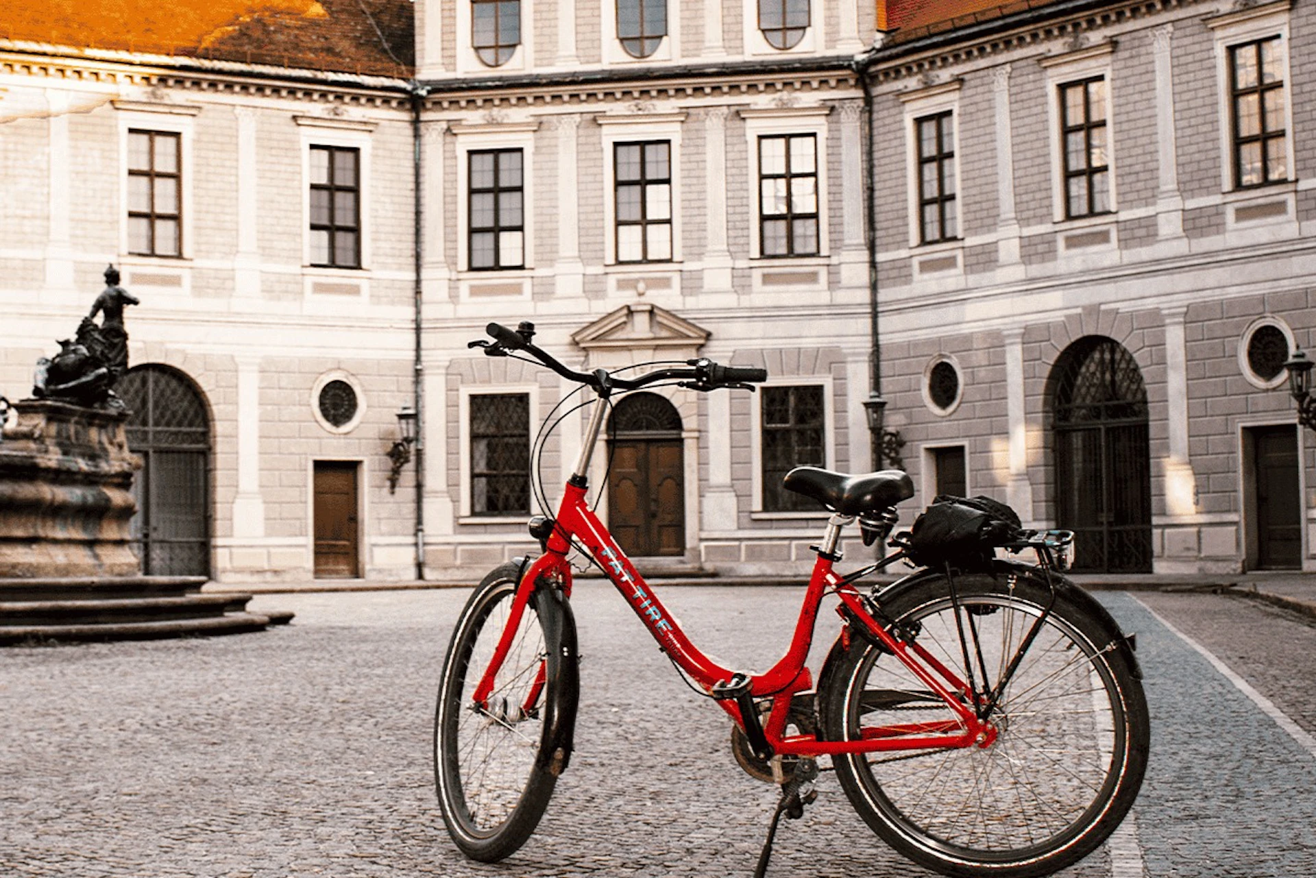 A red bicycle is parked on a cobblestone courtyard in front of a historic building with arched windows and doors.