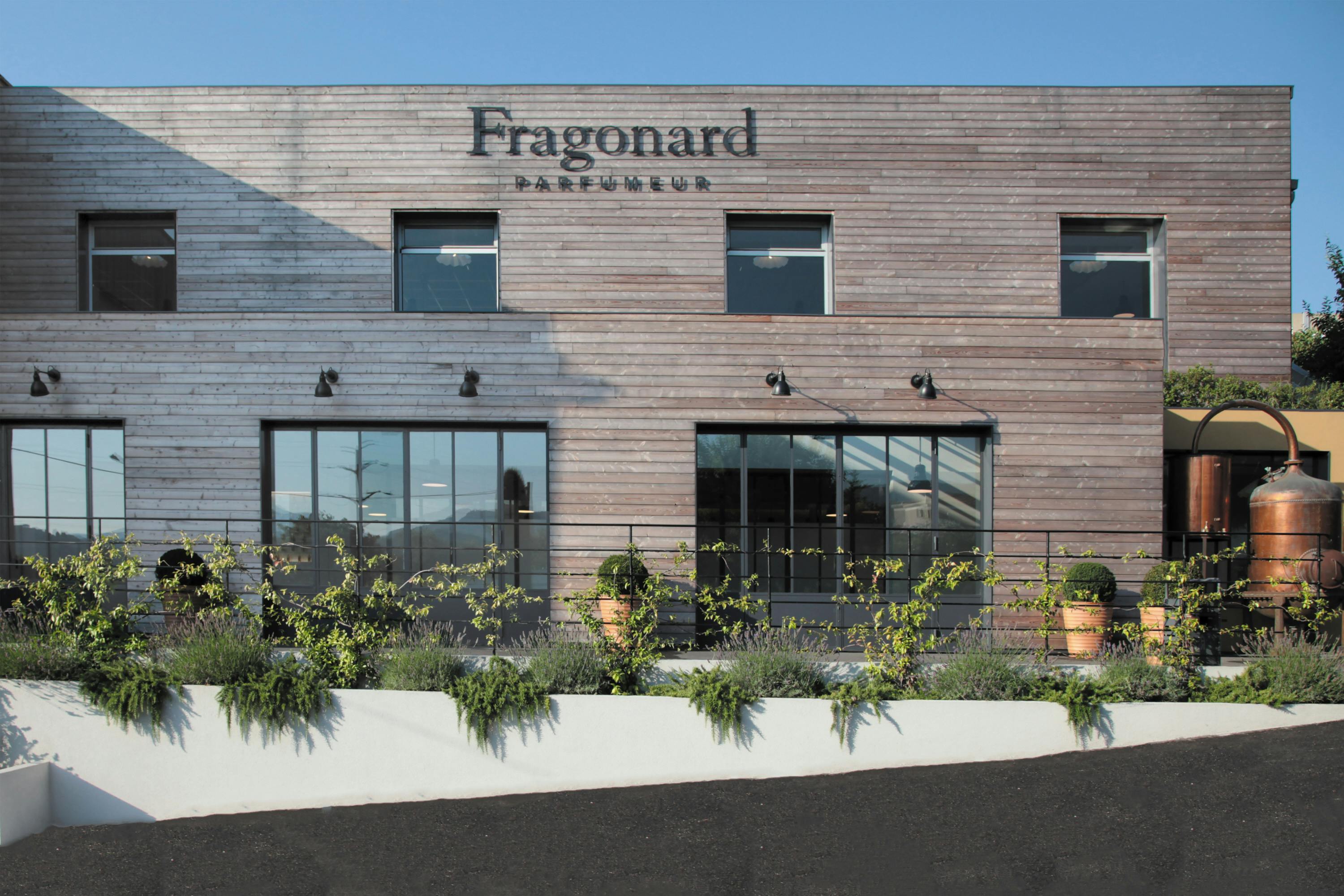 A modern building with "Fragonard Parfumeur" signage, large windows, and a row of potted plants and greenery in front.