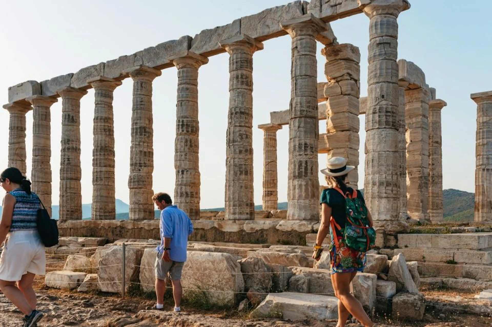 Two people sightseeing near ancient stone columns, with one person wearing a hat and backpack. The sky is clear.