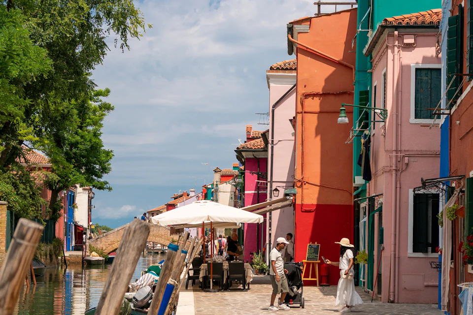 People walking along a canal with colorful buildings, including pink, red, and turquoise walls, under a partly cloudy sky.