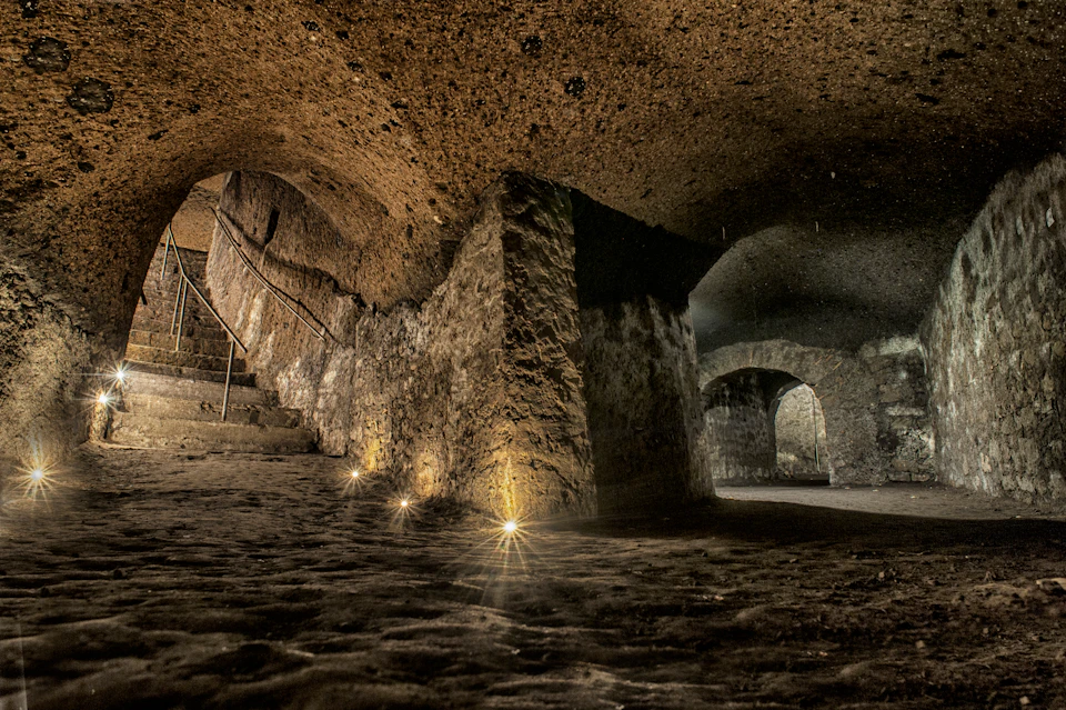 Dimly lit, stone-walled underground tunnel with arched ceilings, steps, and faint lights along the path.