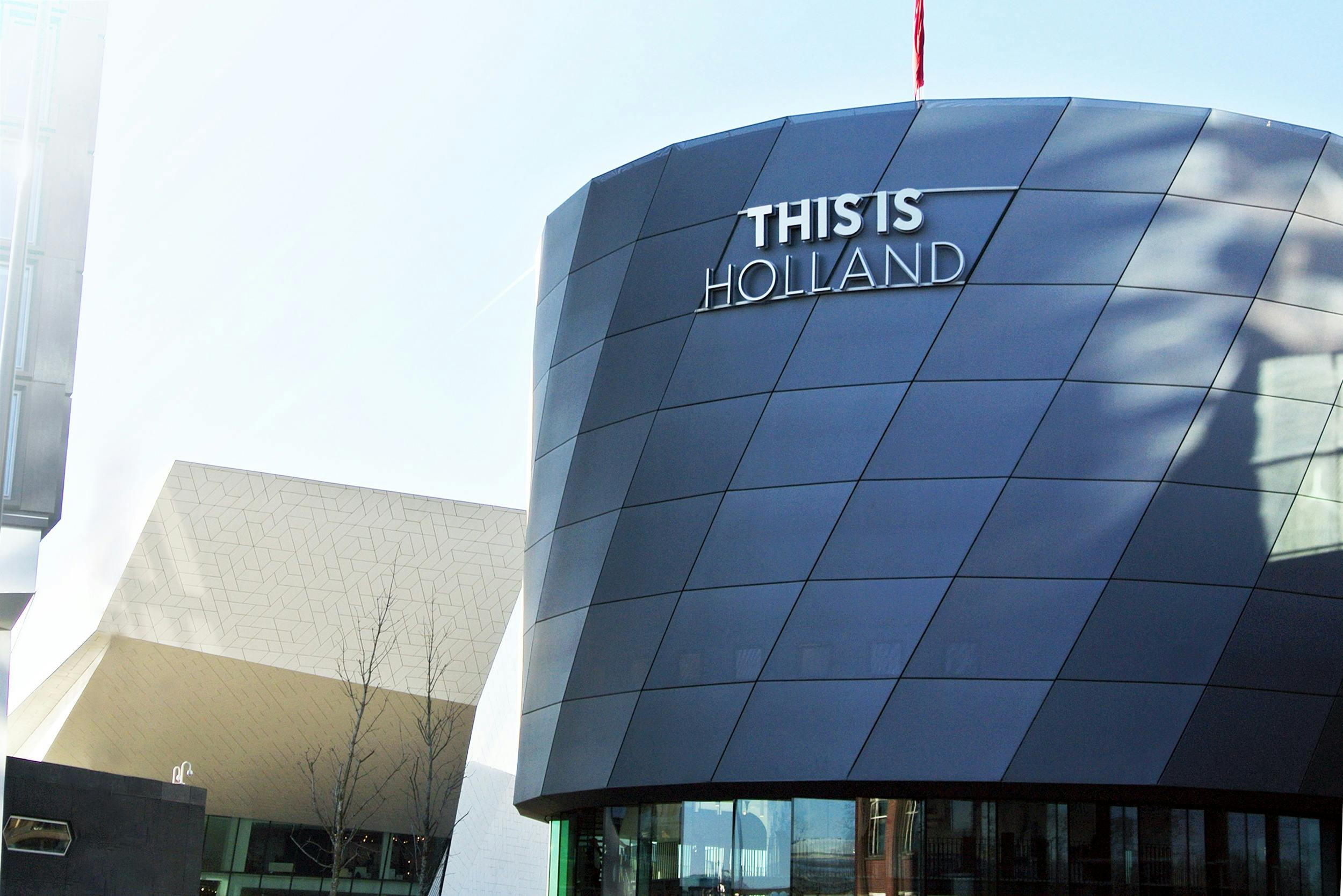 Moderní budova s tmavými geometrickými panely s nápisem „THIS IS HOLLAND“, přiléhající k bílé hranaté stavbě.