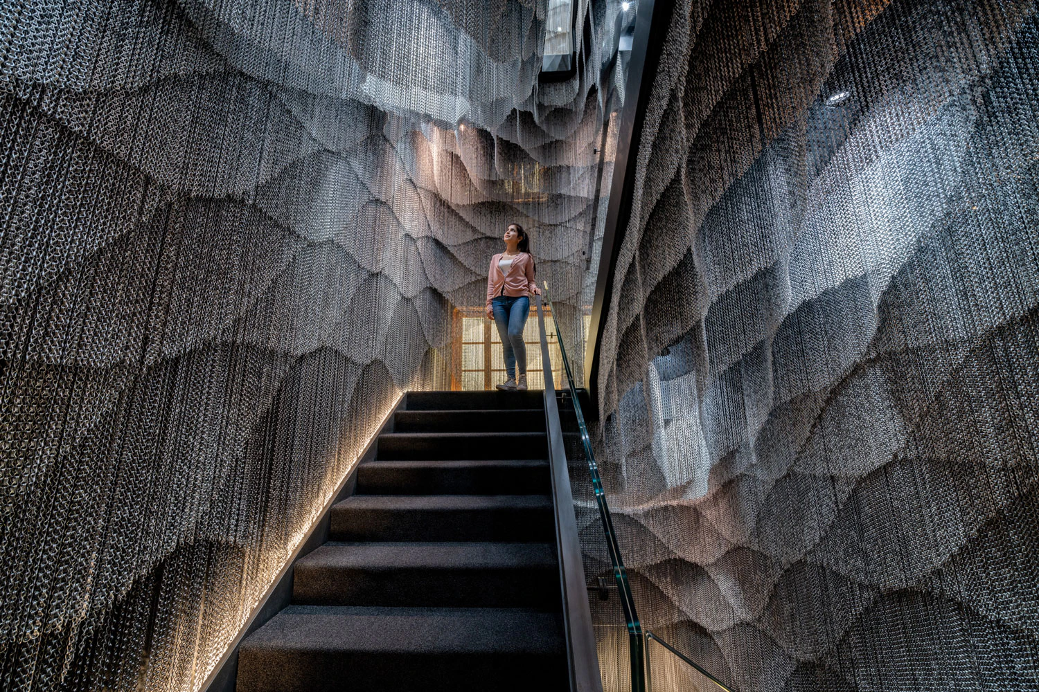 A person stands mid-staircase in a modern, abstractly textured hallway with intricate wall designs and dim lighting.