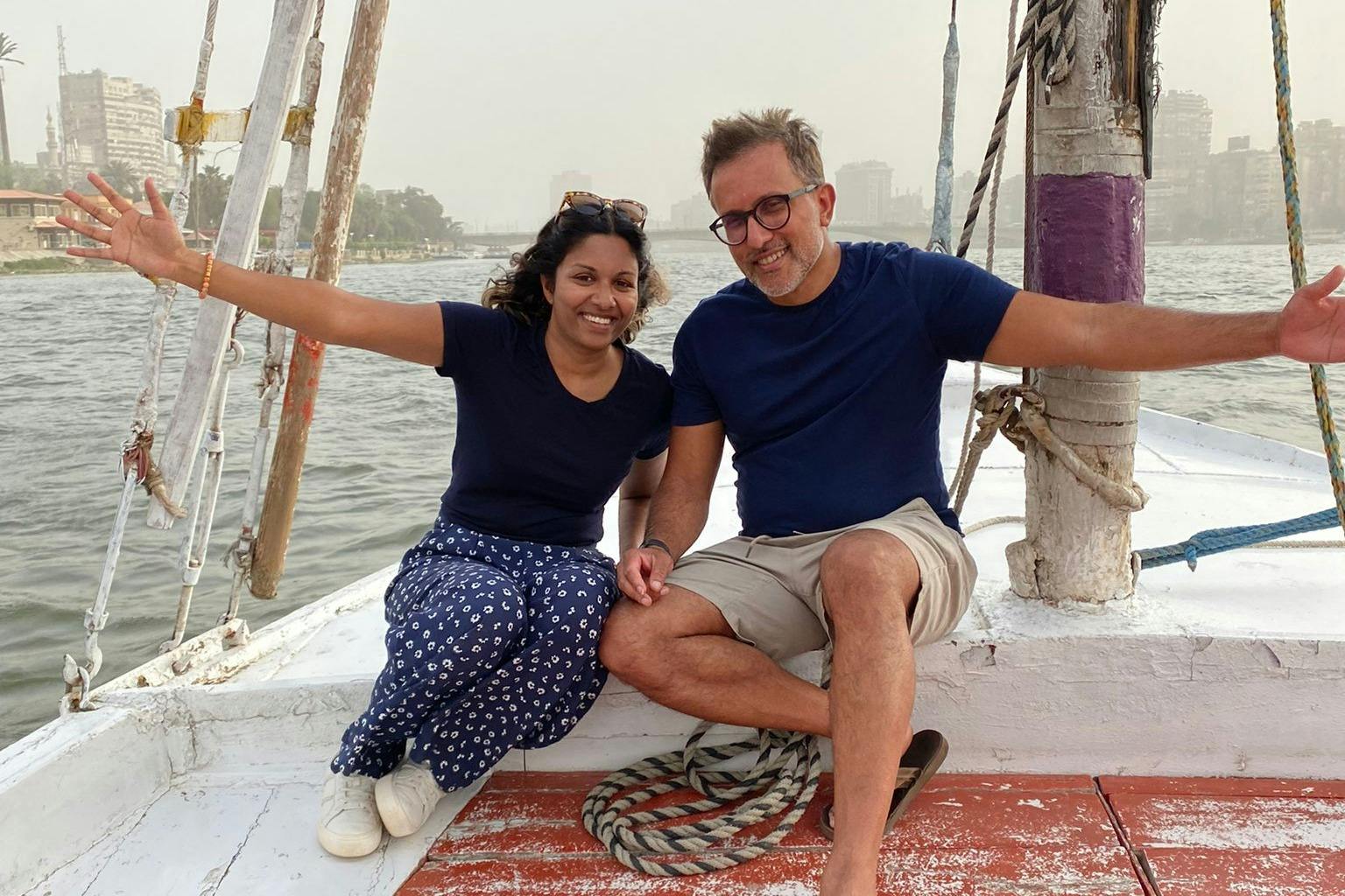 两个人坐在船上，微笑着，背景是水和建筑物，紧紧抓住绳索保持平衡。