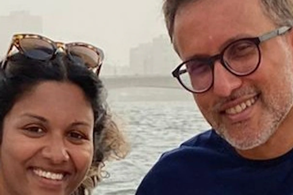 Two people sitting on a boat, smiling, with water and buildings in the background, holding onto ropes for balance.