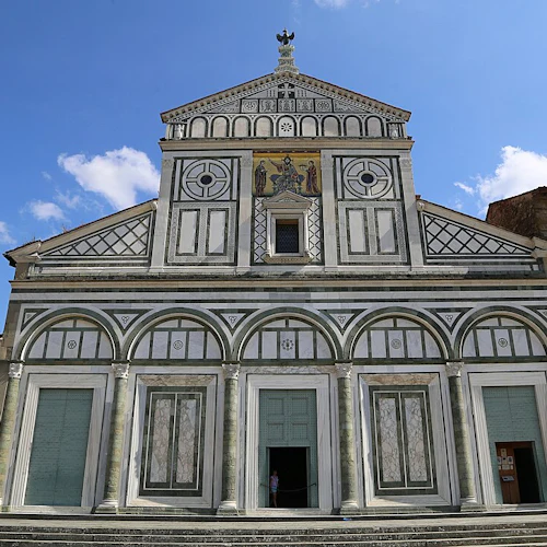 Fasaden på en historisk kyrka med geometriska mönster, bågar och en central mosaik under en klarblå himmel.