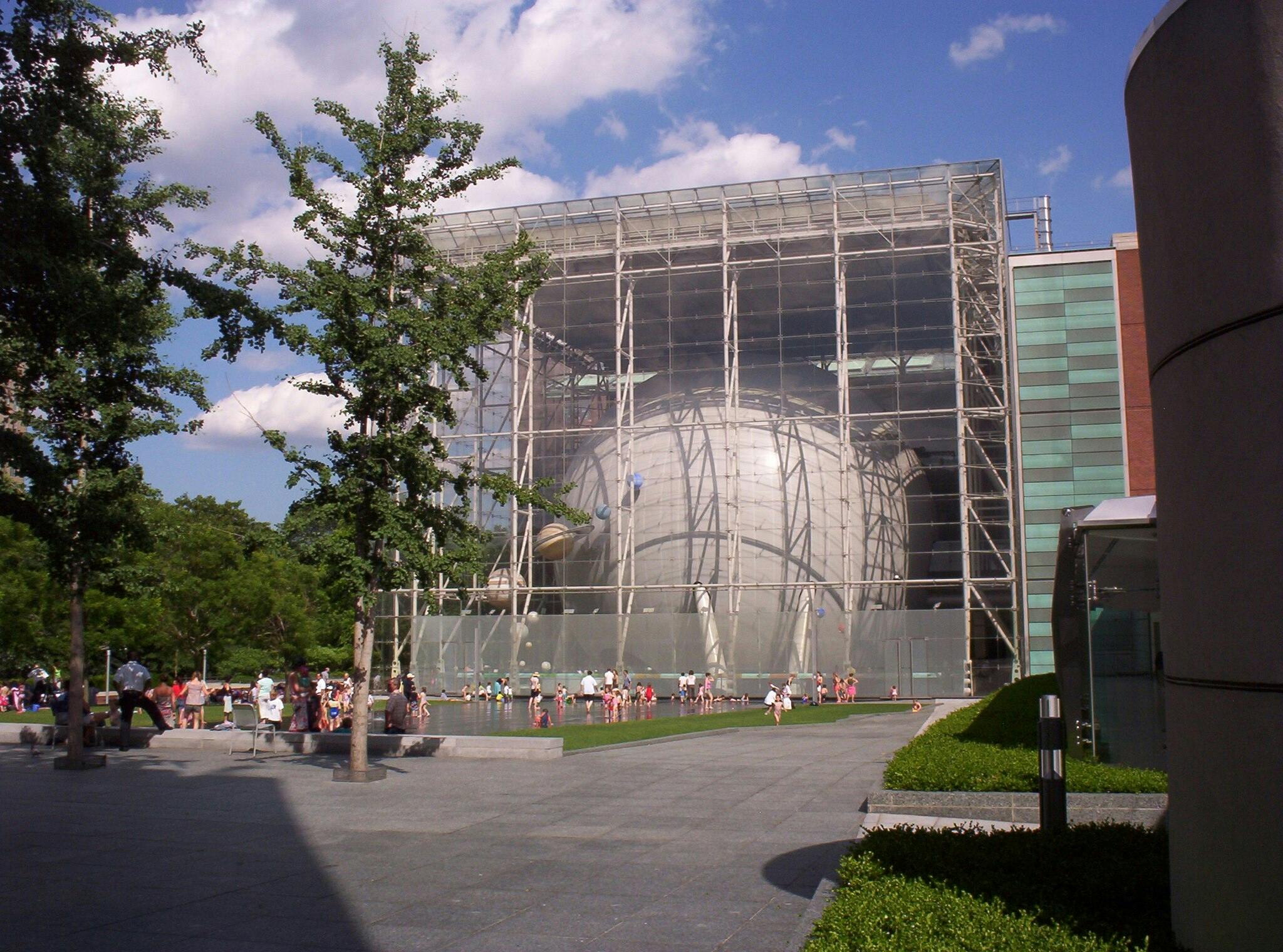 A large glass-fronted building with a spherical structure inside, people gathered on a paved area and grassy lawn outside, trees, and a blue sky.