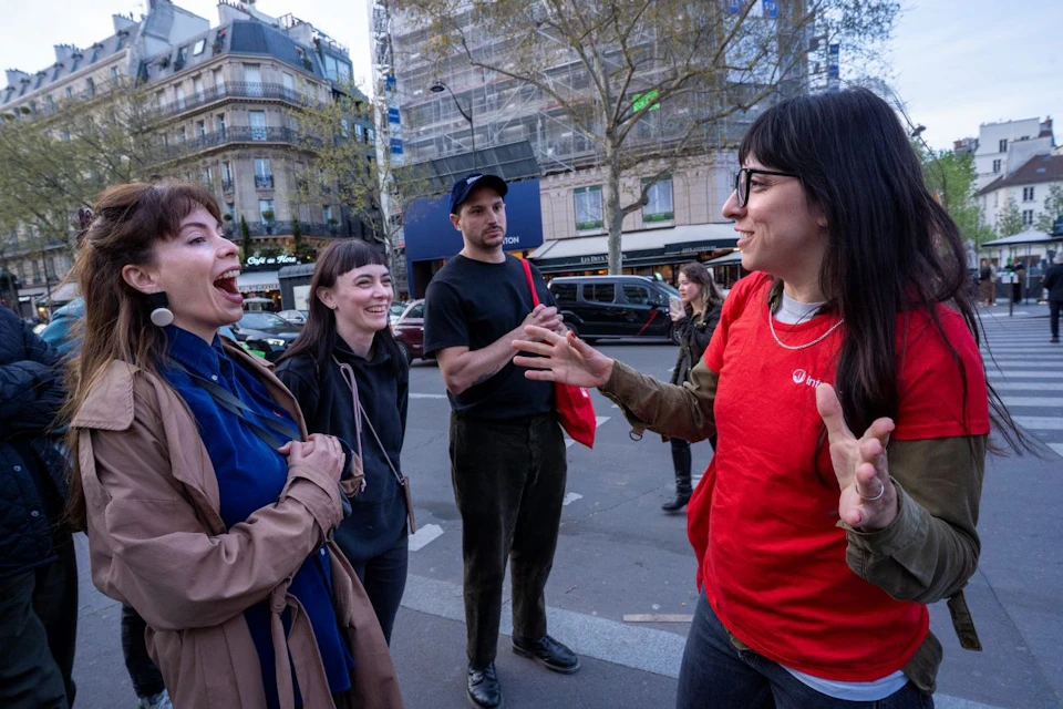 People gathered in a city street, conversing and laughing, with buildings and trees in the background.