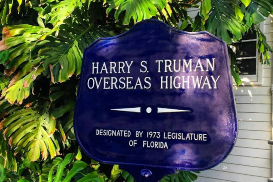 A blue sign reads "Harry S. Truman Overseas Highway, designated by 1973 Legislature of Florida," set against lush green foliage.