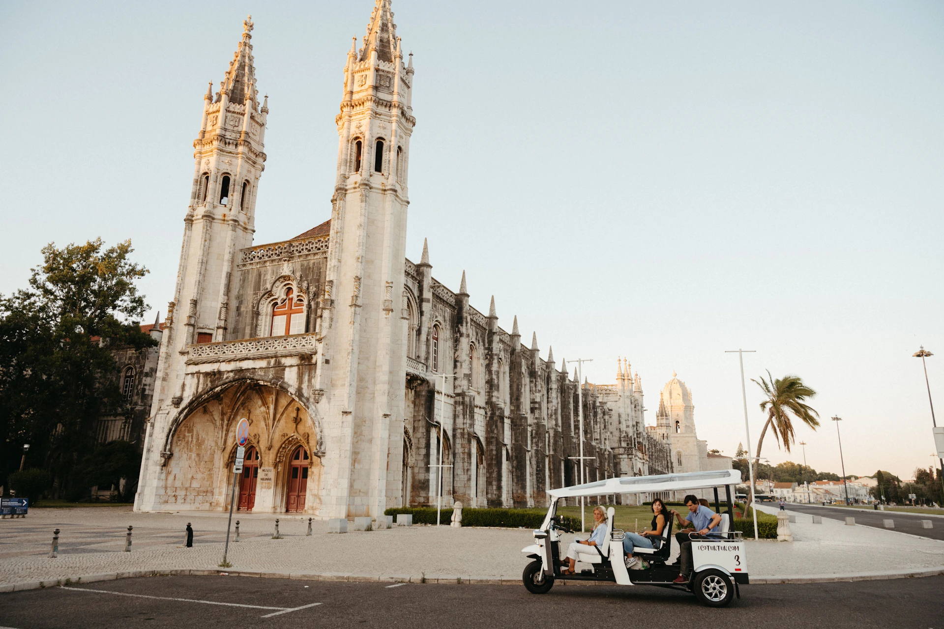 People riding a white tour vehicle in front of a historic stone building with twin towers and ornate gothic architecture.