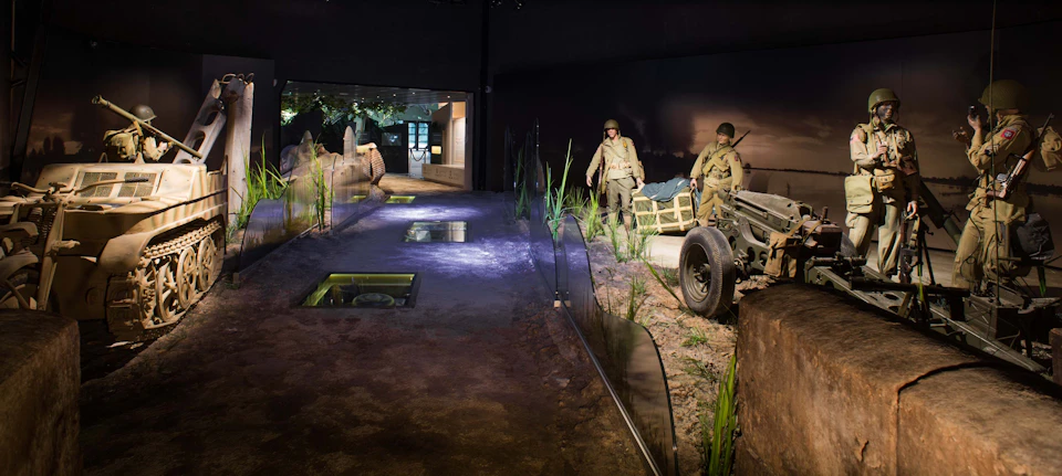 A museum exhibition featuring Second World War soldiers with their equipment and a jeep, set against a realistic battlefield backdrop.