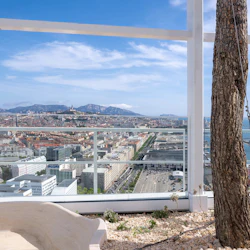 Utsikt över staden från en terrass med träd och växter, med byggnader, berg och havet under en blå himmel.