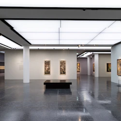 Rymligt, modernt konstgalleri med inramade målningar på vita väggar, blanka golv och taklampor. En svart bänk sitter i mitten.