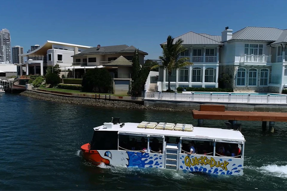 A colorful tour boat labeled "Quack'r Duck" cruises past luxurious waterfront homes on a sunny day.