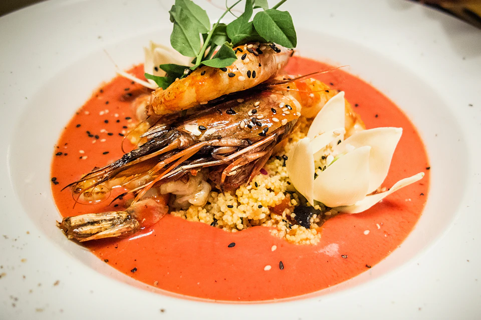 A gourmet dish featuring prawns on a bed of couscous, garnished with greens and seeds, in a red sauce with white vegetable slices.