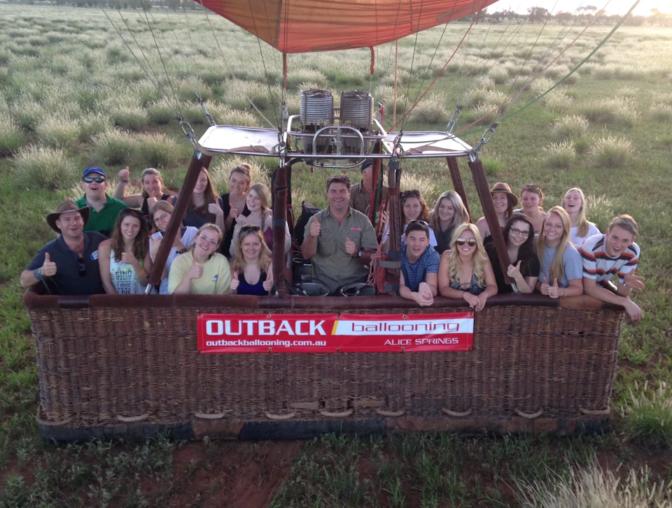 A group of people giving thumbs up in a hot air balloon basket labeled "Outback Ballooning" ready for takeoff over grassy fields.