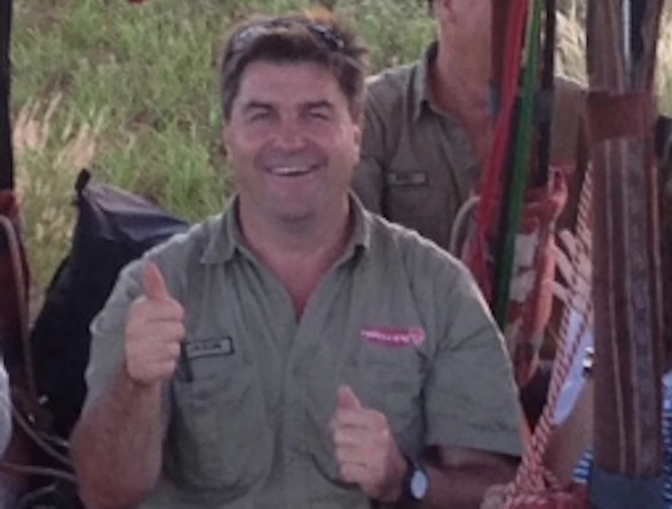 A group of people giving thumbs up in a hot air balloon basket labeled "Outback Ballooning" ready for takeoff over grassy fields.