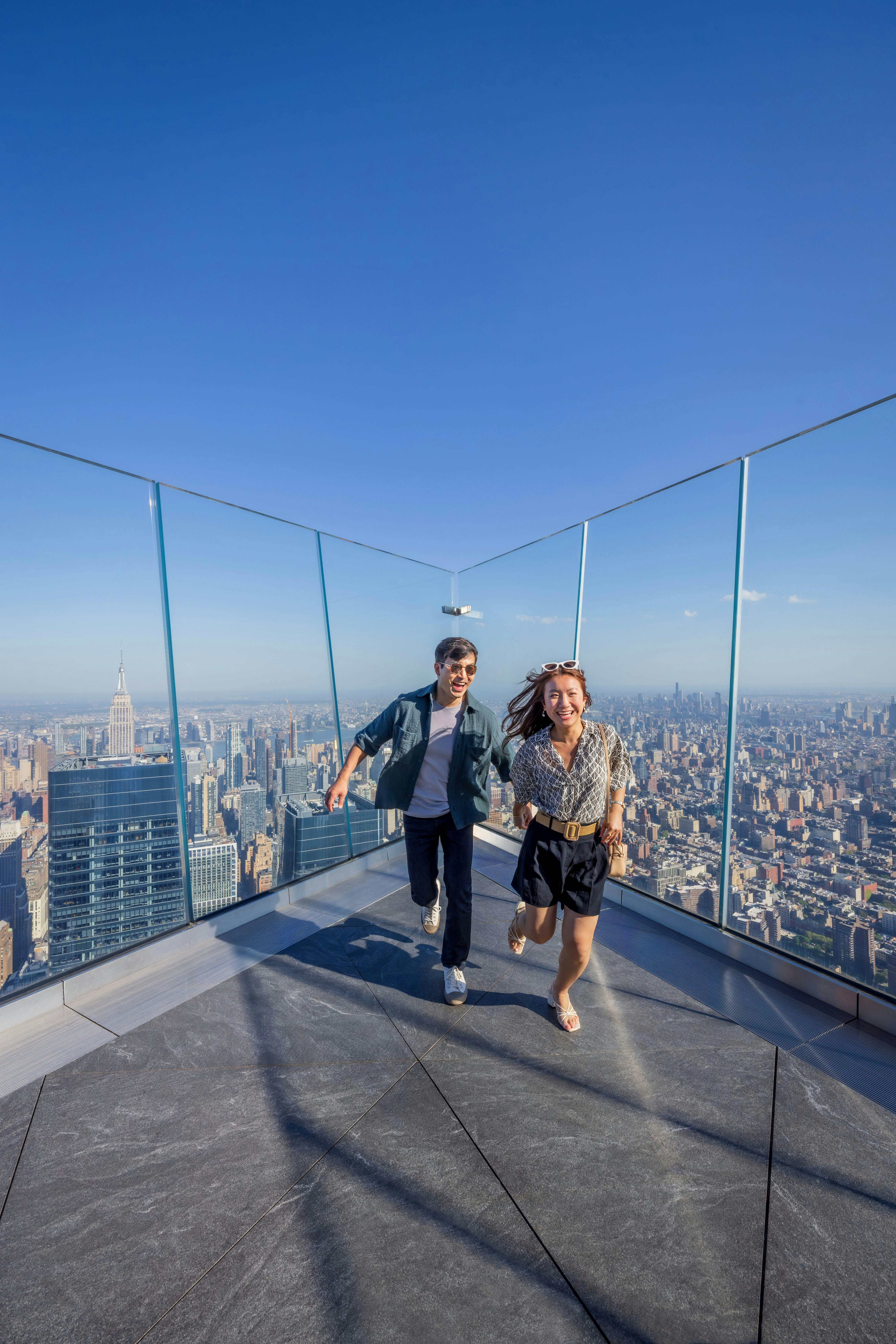 엠파이어 스테이트 빌딩을 배경으로 두 사람이 유리 전망대에서 미소를 짓고 달리고 있습니다.