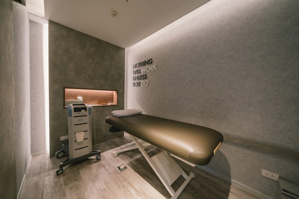 A therapy room with a padded table, a medical device on wheels, and a wall with motivational text reading "Nothing will work unless you do."