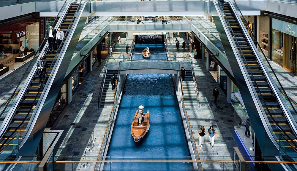 Lidé kráčející po několika patrech velkého nákupního centra s eskalátory a kanálem s loděmi projíždějícími středem.