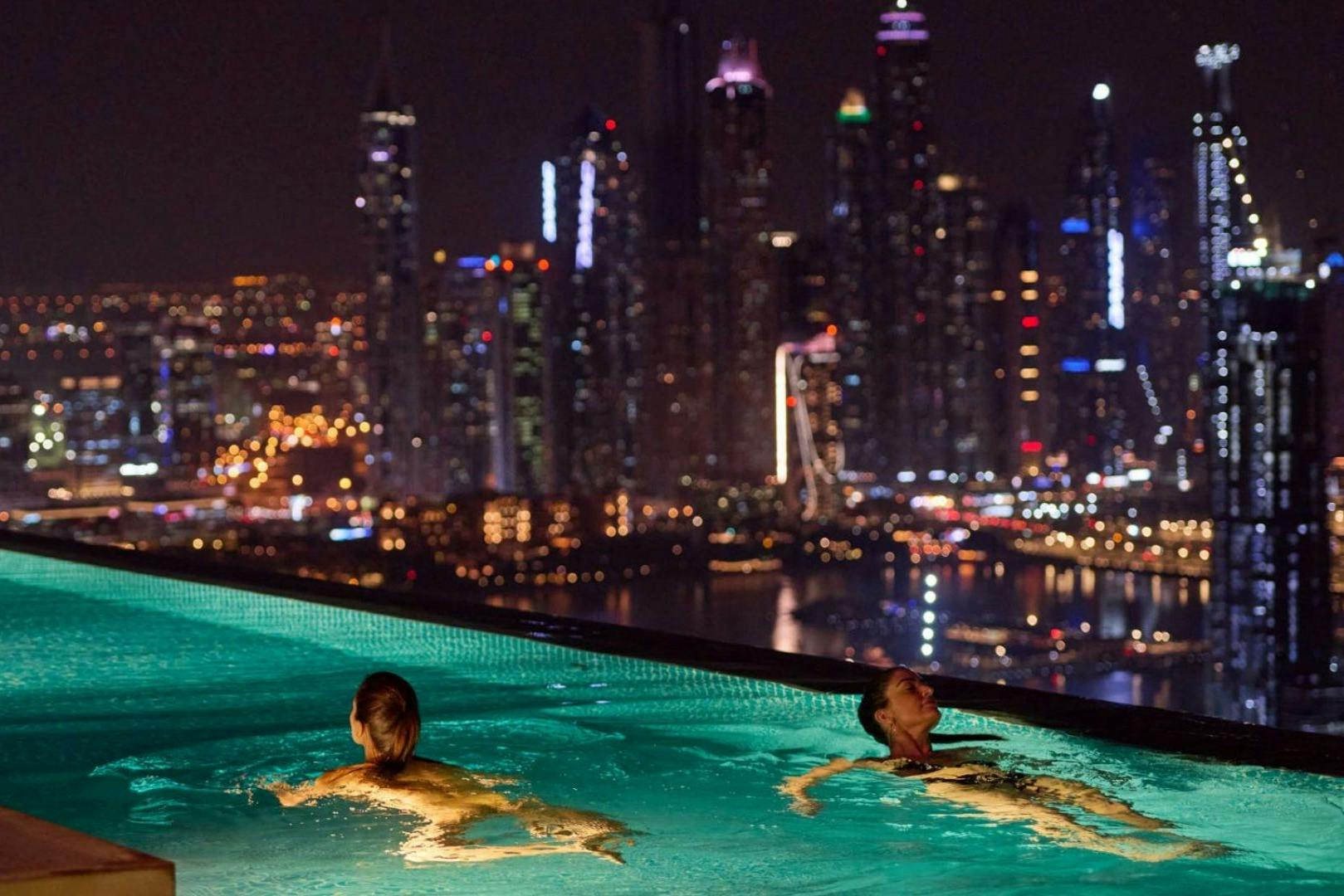 Two people swim in a rooftop pool with a city skyline illuminated by colorful lights in the background at night.