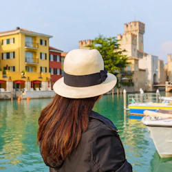A person wearing a straw hat stands by a canal with colorful buildings and a historic castle in the background.