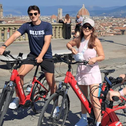 Tre personer står på en utsiktsplats med röda hyrcyklar, leende, med en stadsbild och berg i bakgrunden.