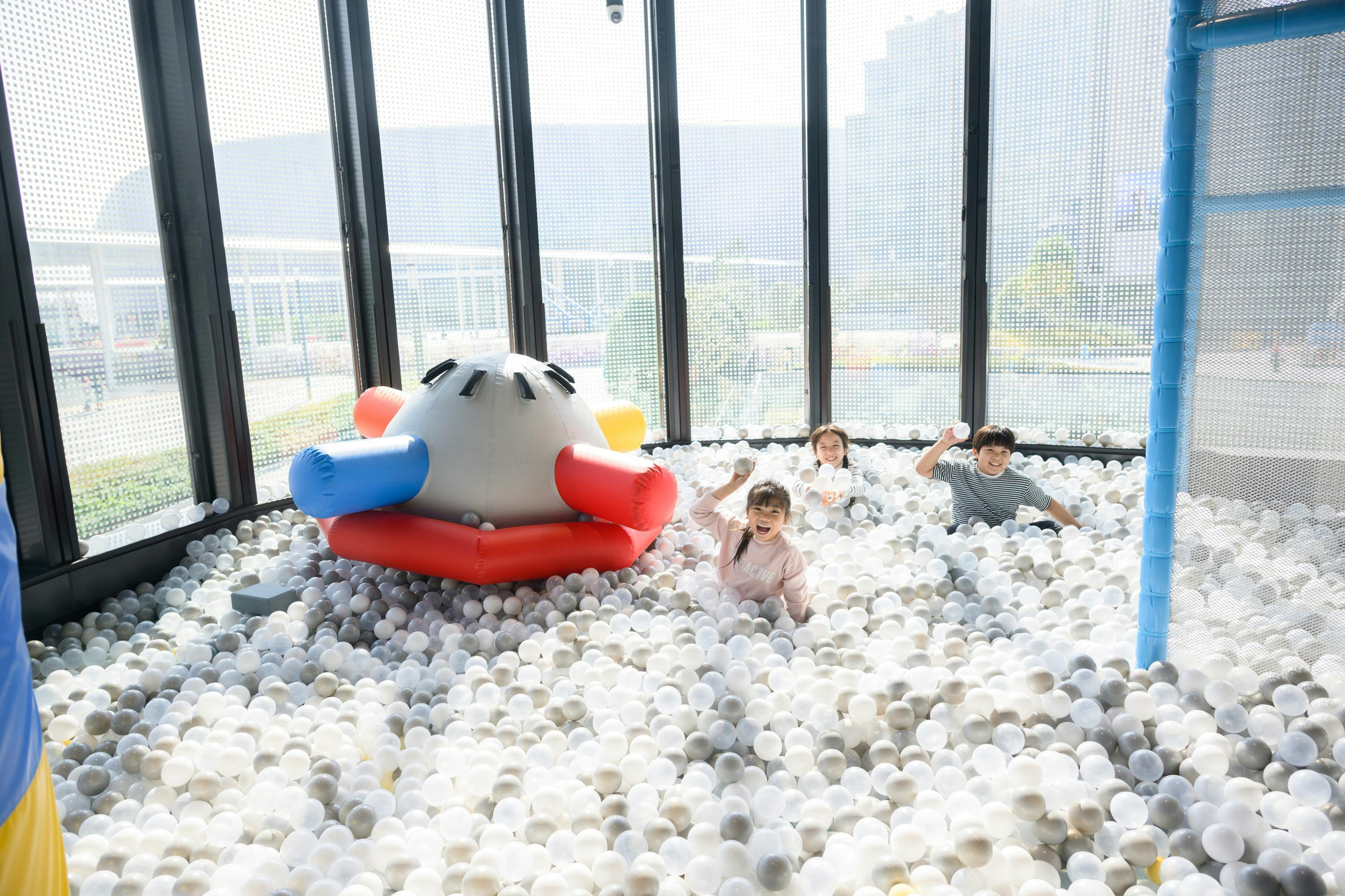 Three children playing in a ball pit filled with white balls, next to an inflatable structure in a bright indoor space.