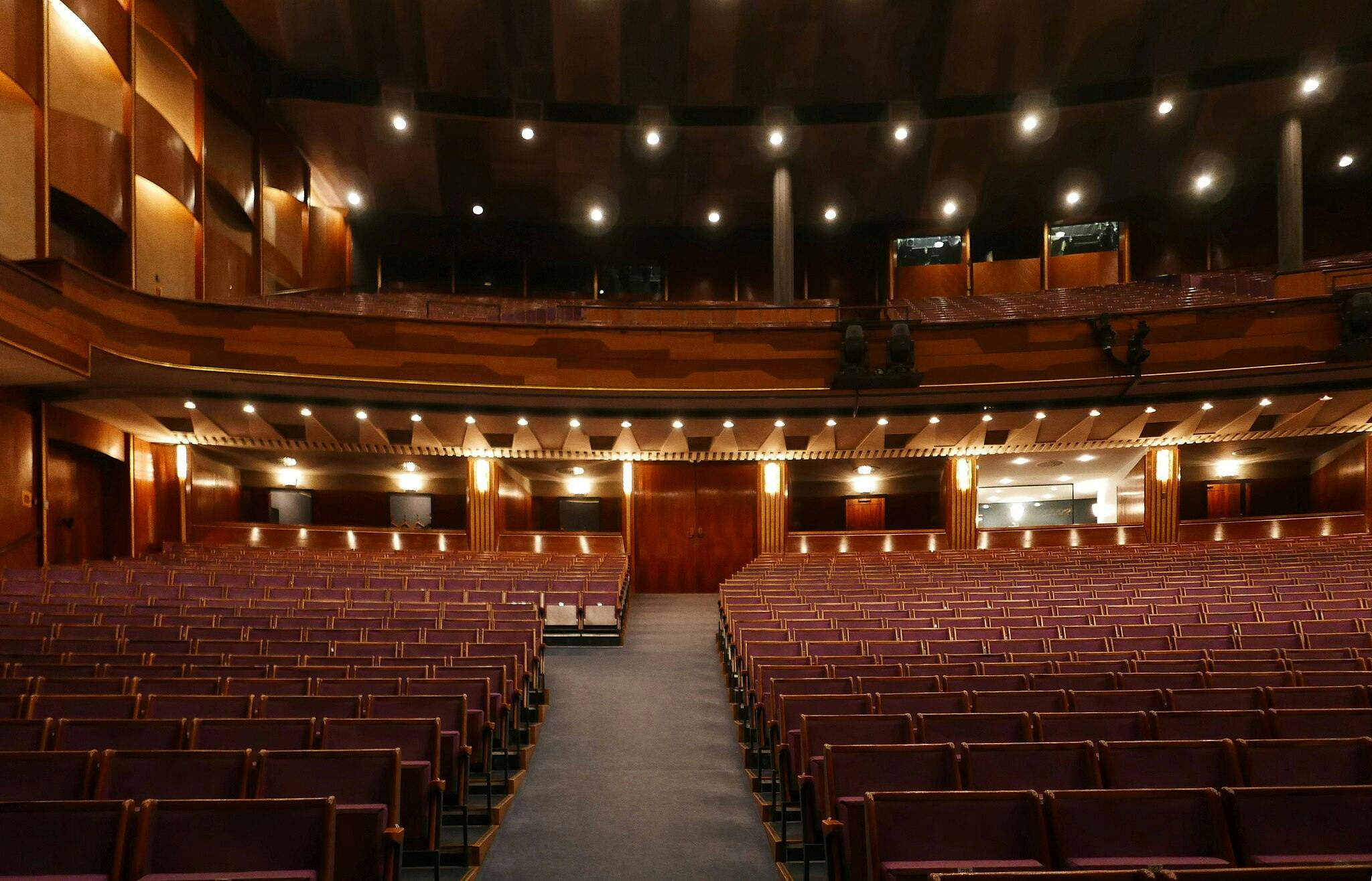 Een lege theaterzaal met paarse stoelen, houten lambrisering en plafondverlichting. Een gangpad leidt naar het podium.
