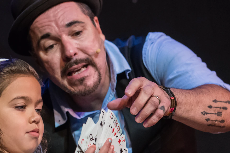 A man in a bowler hat is performing a card trick with a young girl, who is attentively holding playing cards.