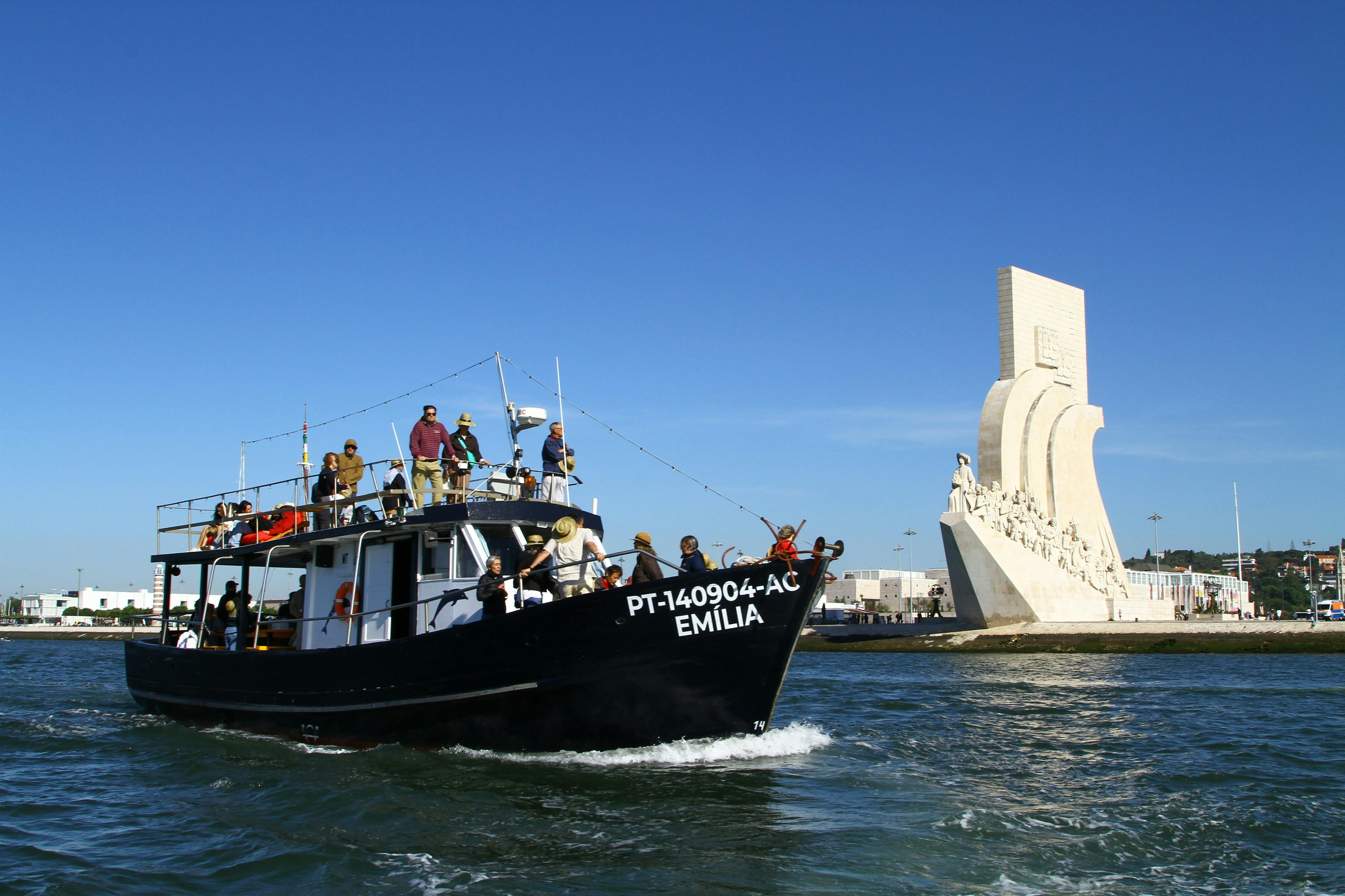 一艘名为“Emília”的黑色船只在水面上，船上有几个人，旁边是一个晴朗的蓝天下的大型海洋纪念碑。