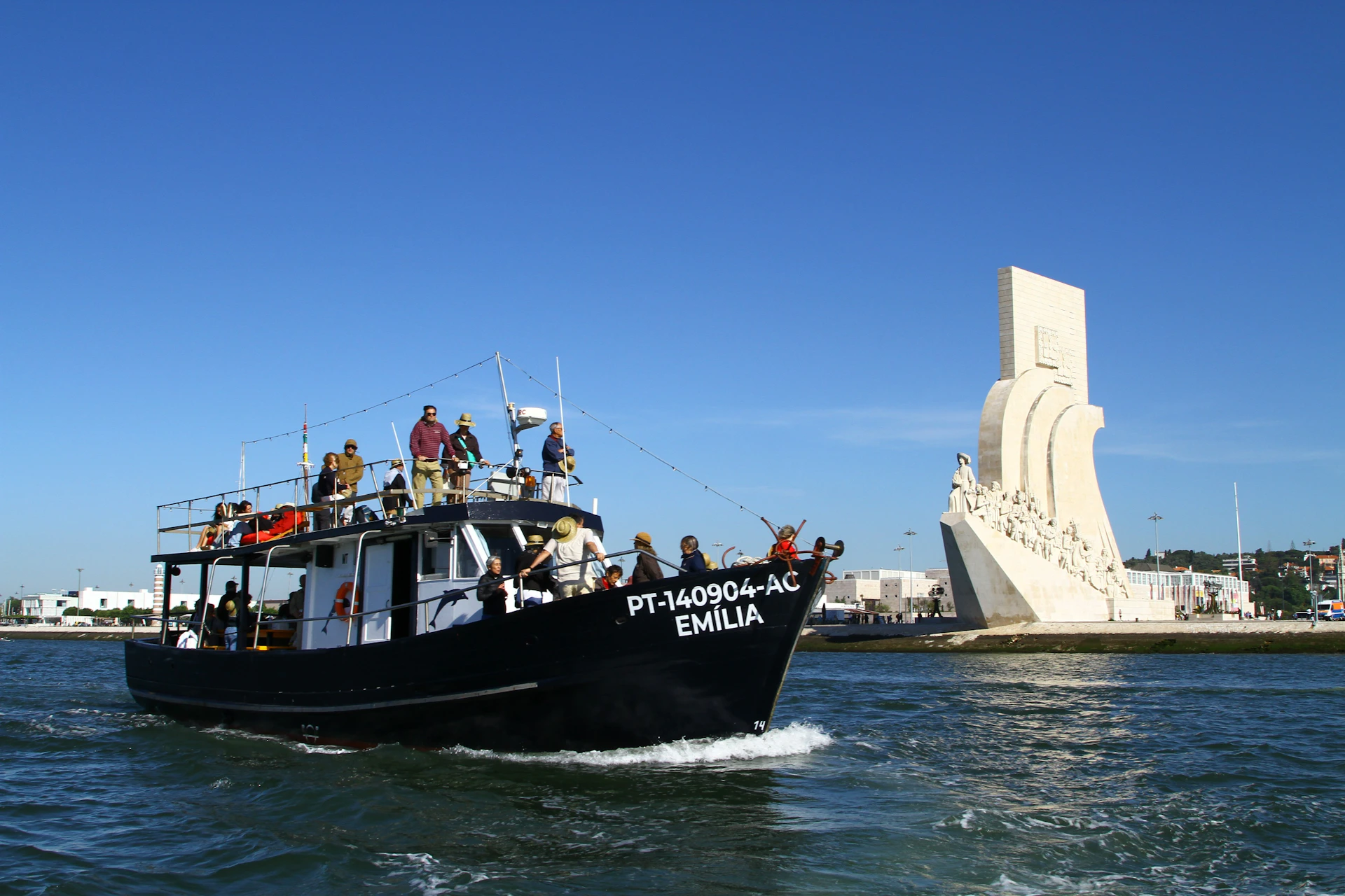 A black boat named "Emília" on water with several people aboard, next to a large maritime monument under a clear blue sky.
