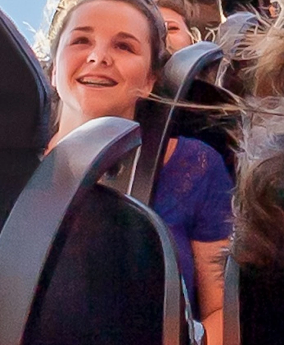 People on a rollercoaster ride scream and laugh with excitement under a clear blue sky with scattered clouds.