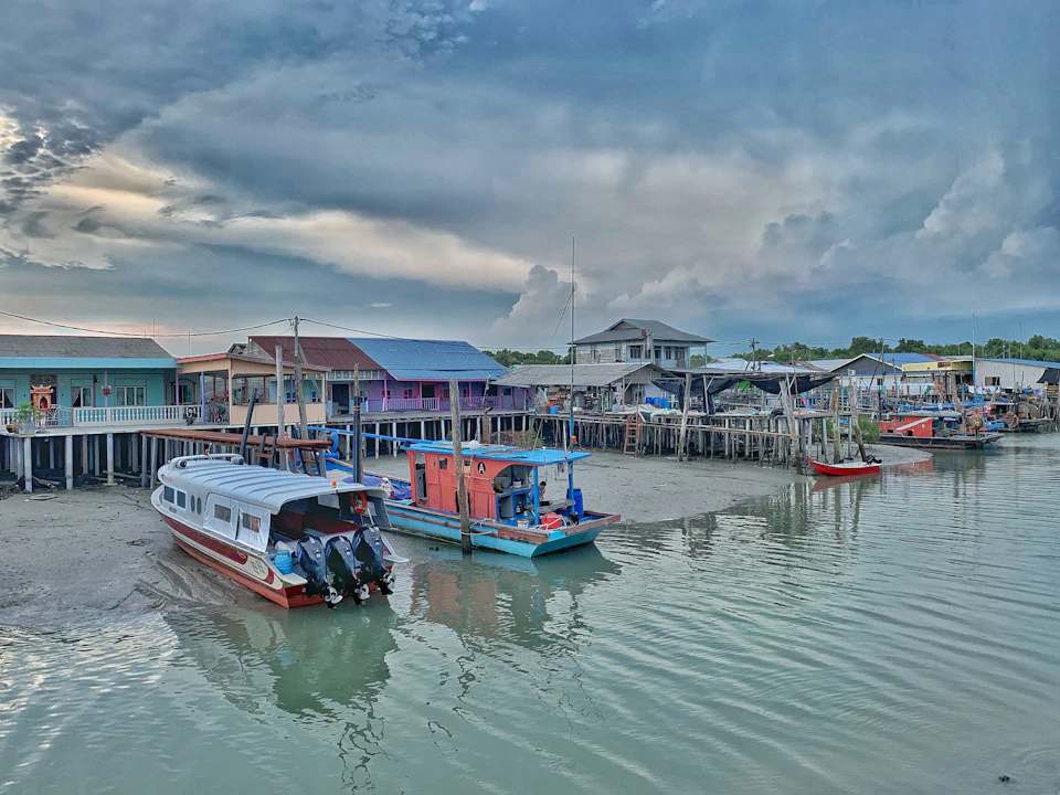Colorful wooden houses on stilts above water with several small boats docked at a quaint riverside village under a cloudy sky.